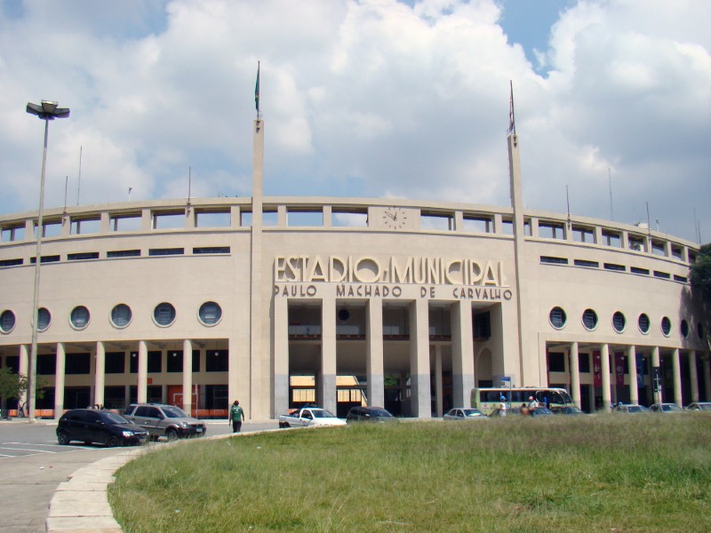 Fachada do Estádio Municipal Pacaembu, com suas colunas e anel de arquibancada. Há alguns carros estacionados em frente